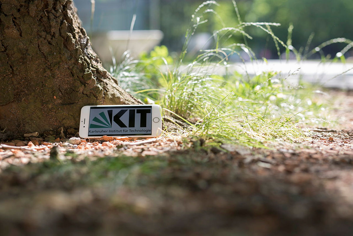 Ein Handy lehnt an einen Baum, der von Gras umgeben ist. Auf dem Bildschirm ist das KIT-Logo abgebildet. 