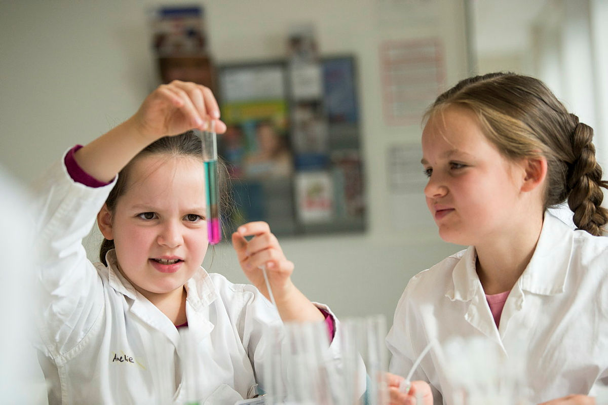 Two kids in white lab coats conduct a science experiment with a dropper in a classroom.