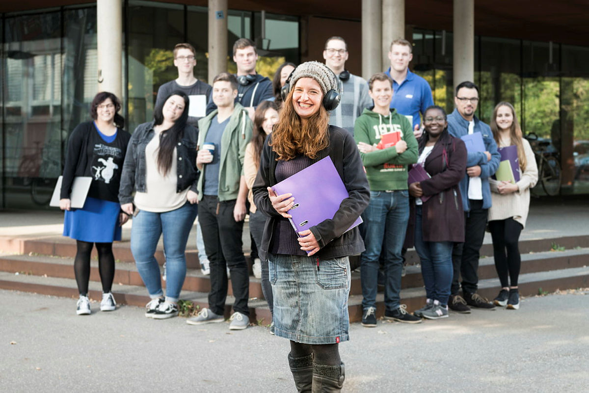 Mehrere Studierende posieren mit Ordnern und Heften in der Hand vor einem Haus. 