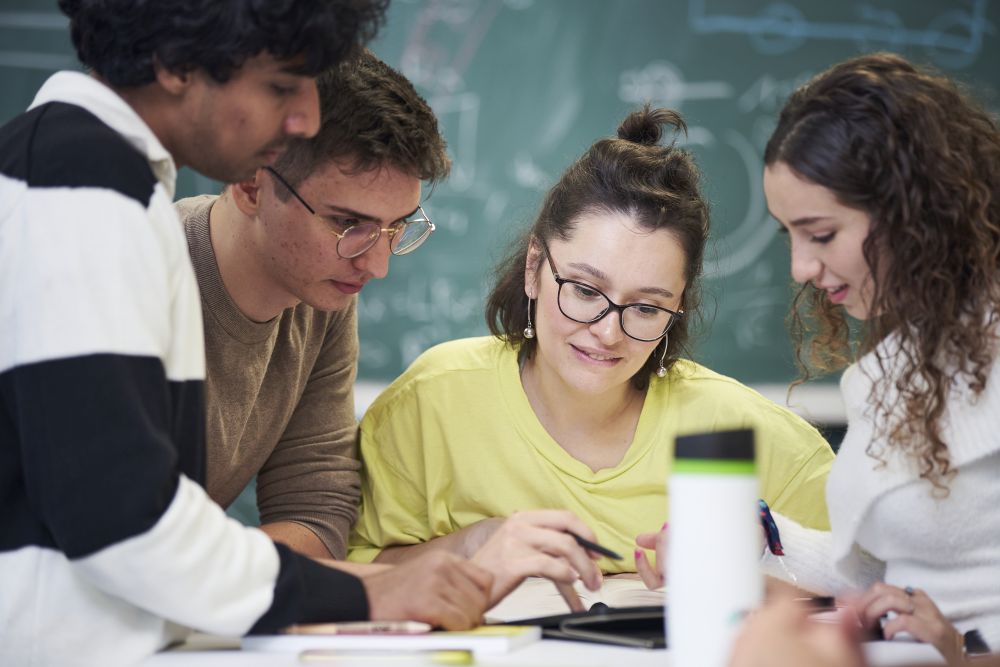 Gruppe junger Studierender arbeiten an Notizen am Tisch vor einer Tafel.