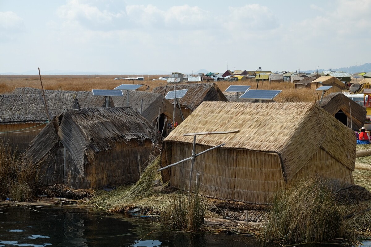 A village of thatched huts stands by a body of water. Next to each hut is a solar cell.