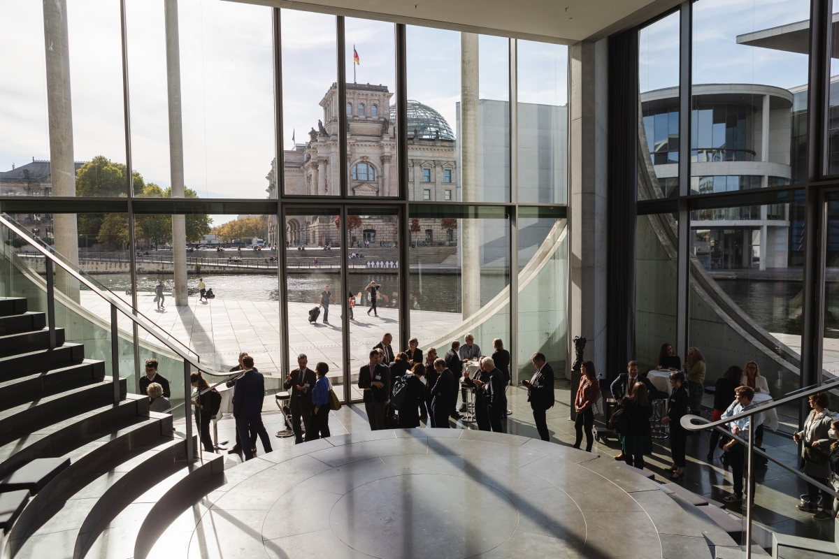 In einem verglasten Foyer versammelt sich eine Menschengruppe. Im Hintergrund ist der Deutsche Bundestag zu erkennen.