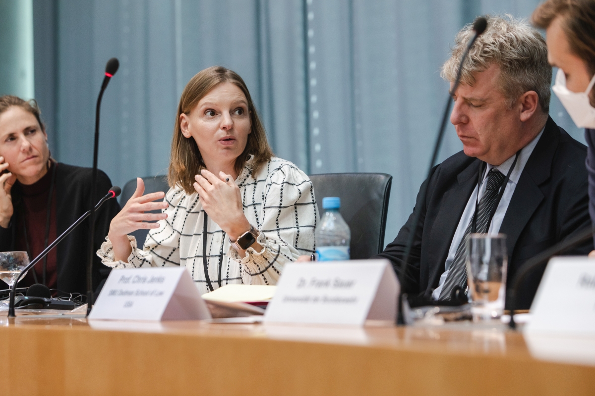 Eine Frau spricht und gestikuliert dabei mit den Händen. Die Personen links und rechts von ihr hören konzentriert zu. Der Mann rechts neben ihr macht dabei Notizen.