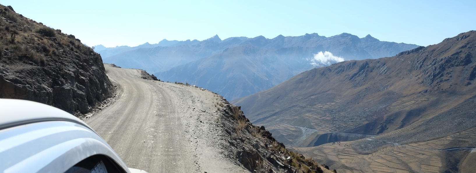 A car drives along a gravel road on a steep slope. In the background: a mountain panorama.