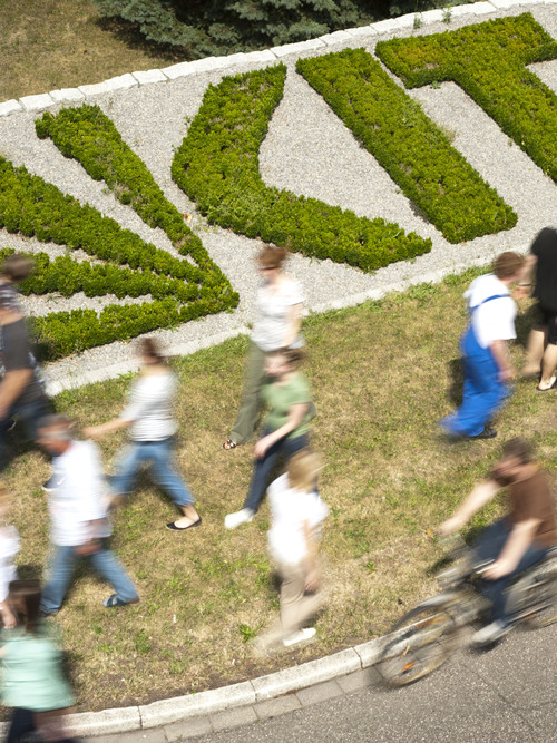 Blurred people walking on grass beside hedge shaped like letters, with one person cycling.