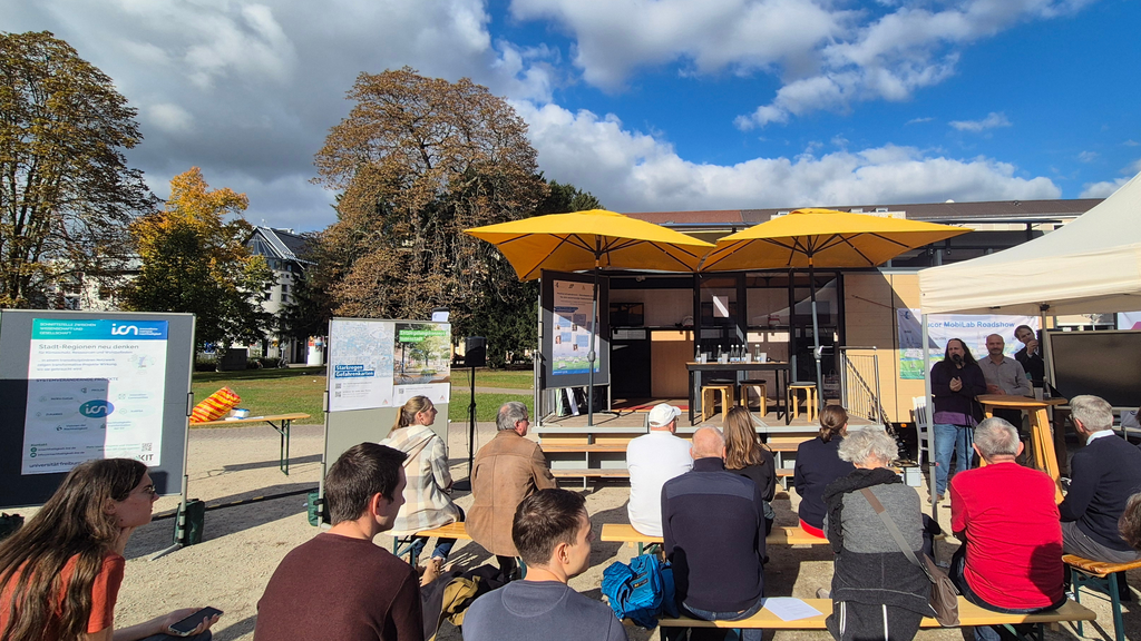 Menschen sitzen vor einer Bühne mit Plakaten an der Seite und schönem Wetter