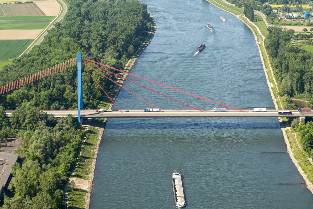 Luftaufnahme der Rheinbrücke bei Speyer. Über die Brücke fahren Autos und Lastwagen, auf dem Fluss verkehren Frachtschiffe.