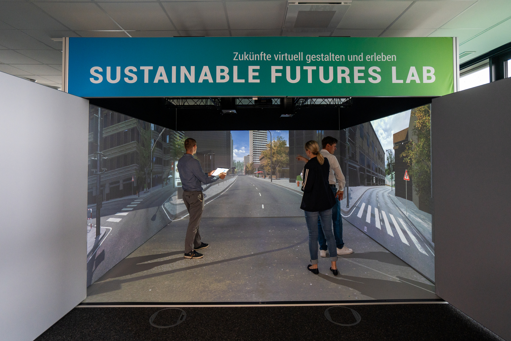 Three spectators standing in a room with city images projected on the walls. The room is titled "Sustainable Futures Lab"