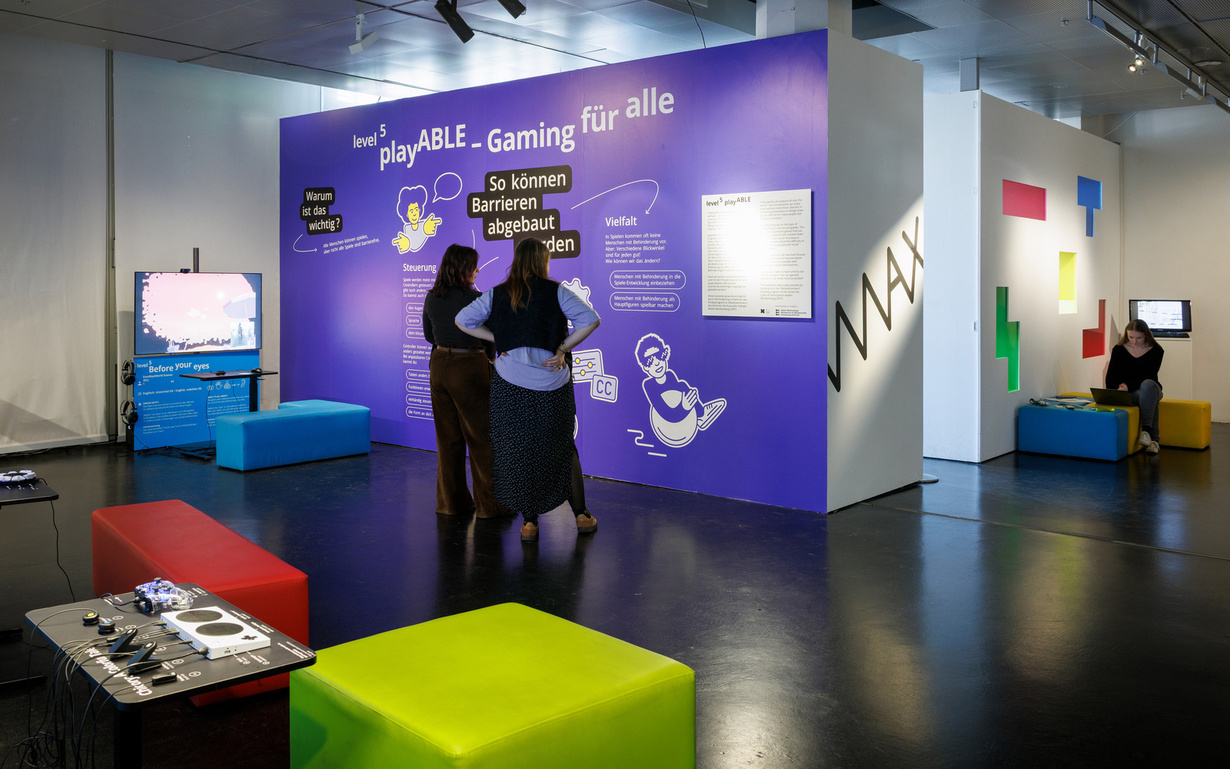 Two people look at a wall whit the writing "playABLE - Gaming für alle. So können Barrieren abgebaut werden"
