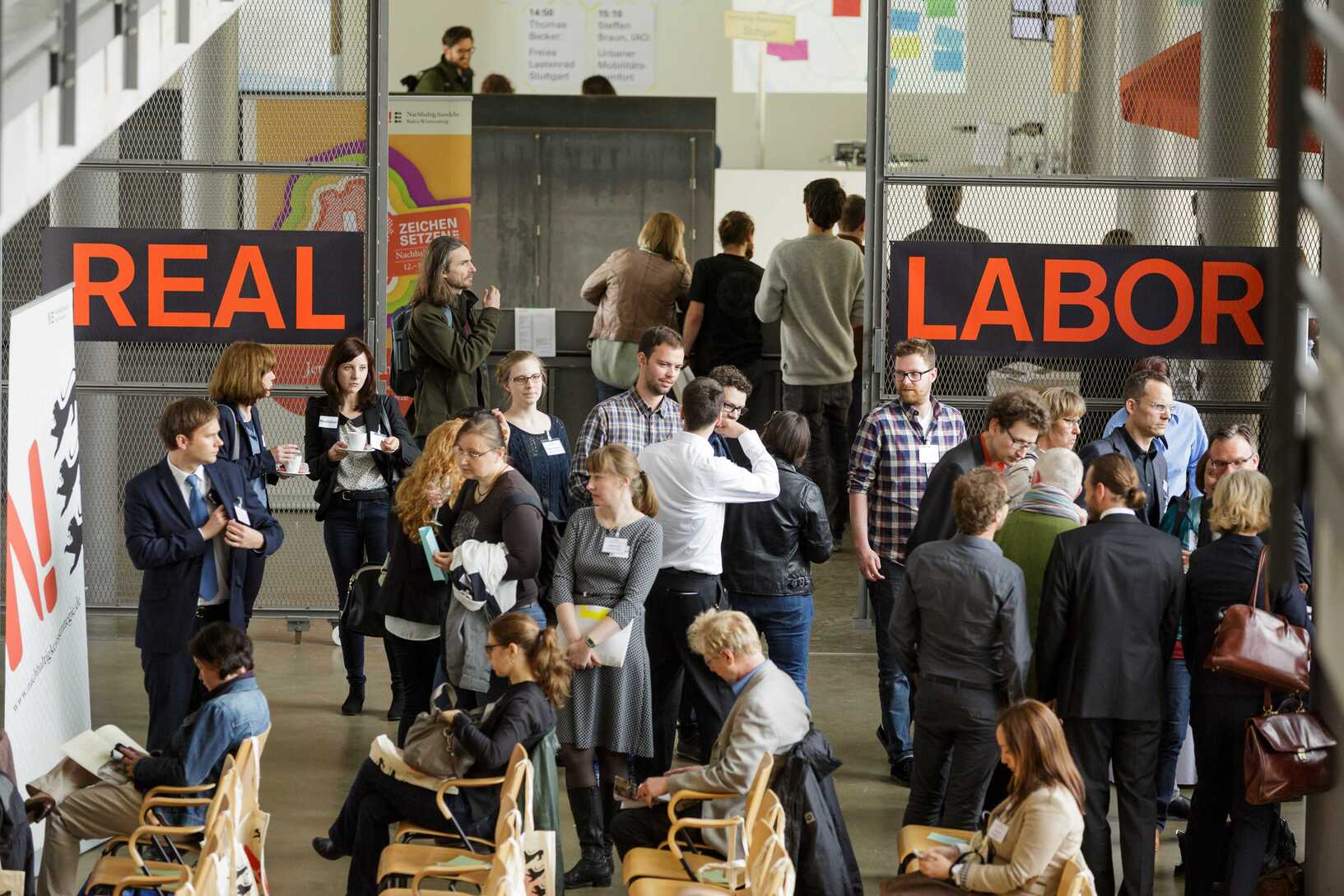 Around twenty people chatting in groups of up to five people. The german words "Real" and "Labor" are spelled on the wall in the background